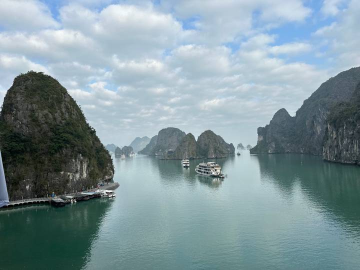 Panoramic view of emerald waters and limestone karst islands of Ha Long Bay dotted with cruise boats beneath partly cloudy skies.
