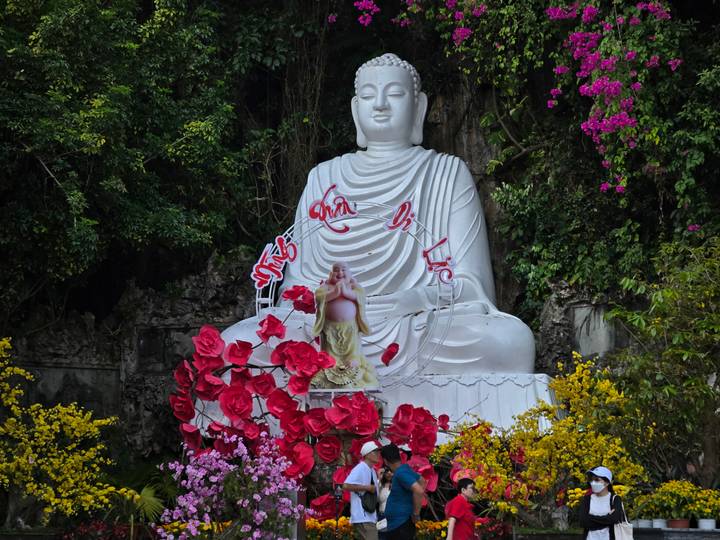 Large white Buddha statue seated outdoors surrounded by bright pink and yellow flowers.