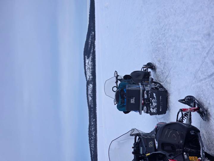 Two snowmobiles parked on a vast frozen lake with forested hills in the distance under a gray sky.