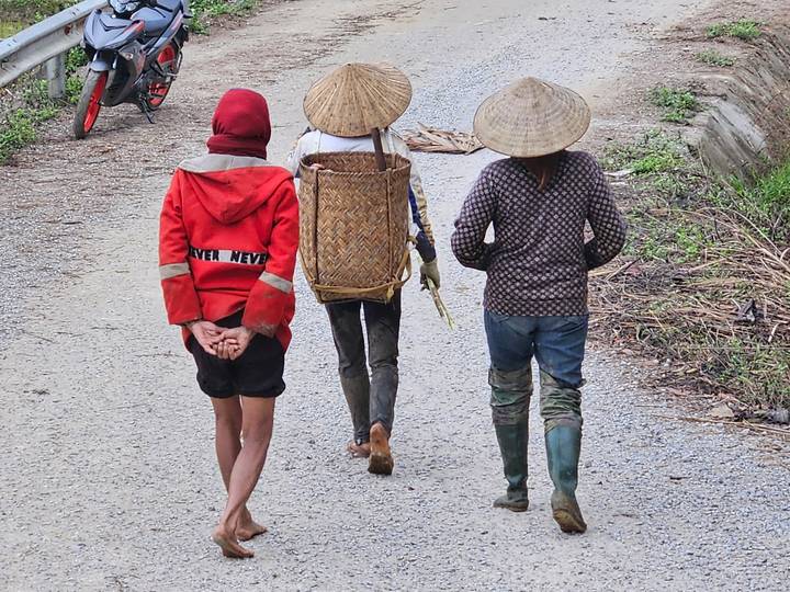 Three local villagers walk along a rural gravel road, one carrying a woven basket backpack