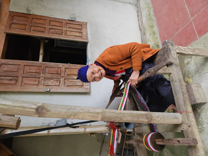 Elderly woman smiling while weaving colourful fabric on a traditional loom in a village house