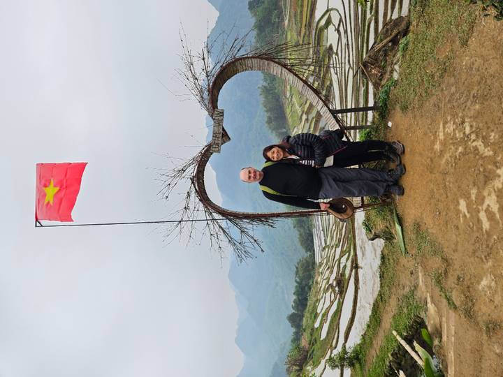Travelling couple pose under a heart-shaped wooden arch with terraced hills and mountains behind
