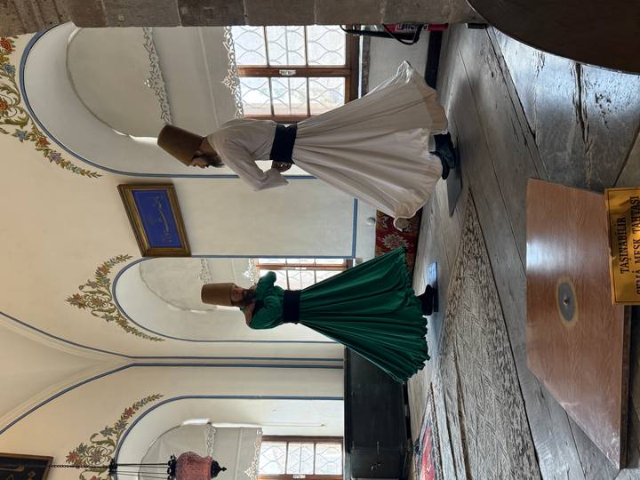 Two whirling dervishes in green and white robes perform inside a historic hall.