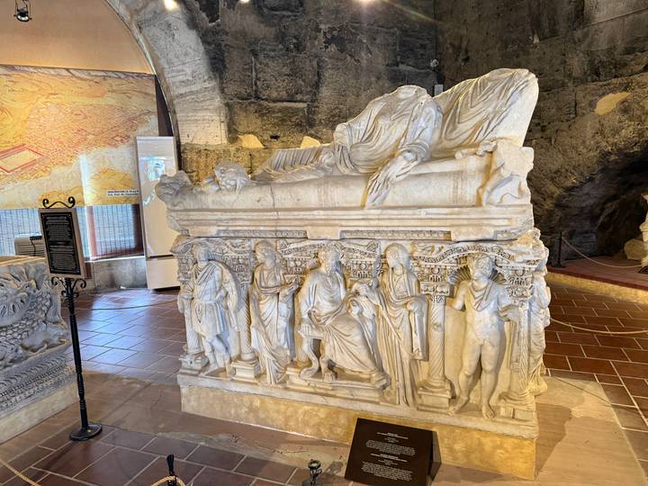 Marble sarcophagus with detailed reliefs displayed in a small museum room.