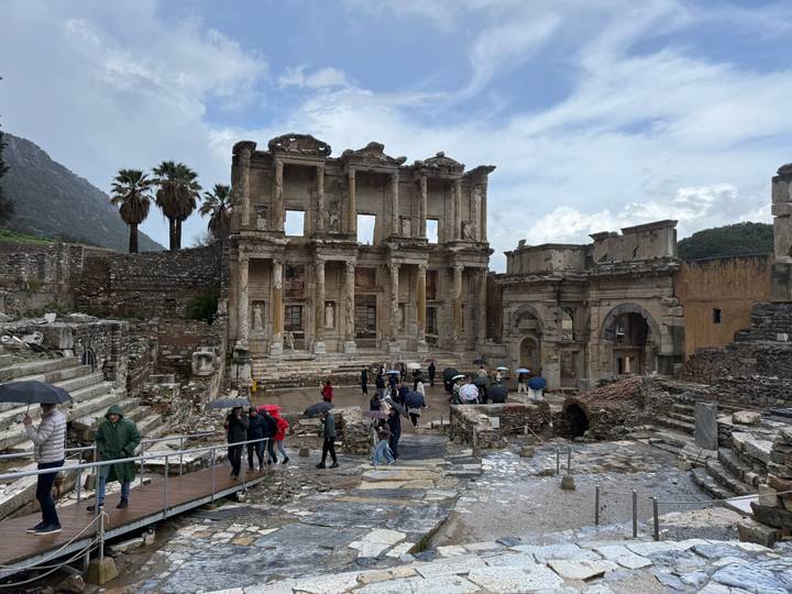The grand facade of the Library of Celsus with visitors walking under umbrellas.