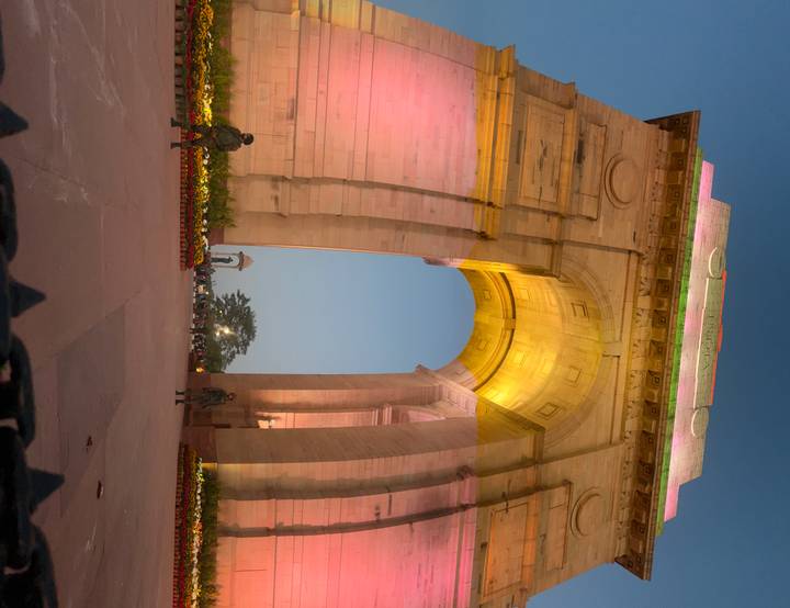 Up-lit arch of India Gate glowing pink and yellow during early evening against a blue sky.
