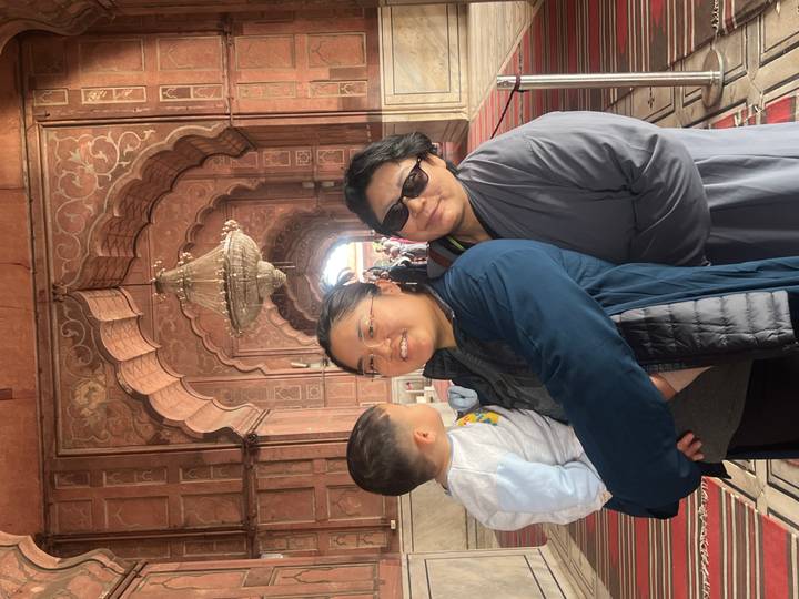Mother, grandmother and toddler pose happily in a Mughal red-stone corridor with chandeliers.