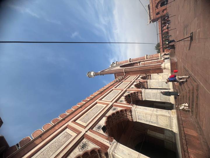 Side façade of Jama Masjid with marble detailing and people climbing steps beneath a clear sky.