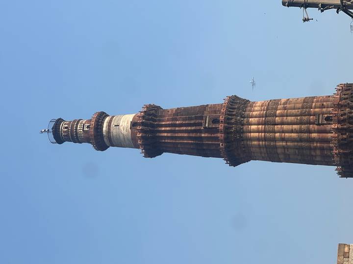 Tall red-sandstone Qutub Minar tower rising against a clear blue Delhi sky with a small airplane in the distance.