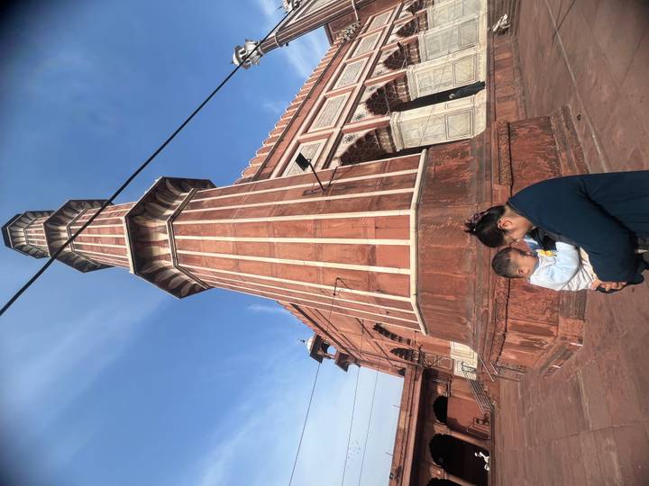 Mother holding a small child at the base of a red-sandstone mosque minaret under a bright blue sky.