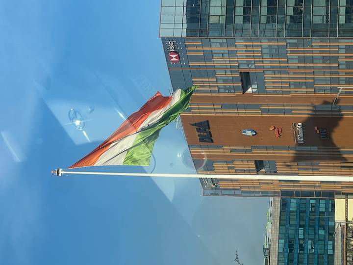 Large Indian tricolour flag waving on a tall pole beside modern glass offices bearing corporate logos.