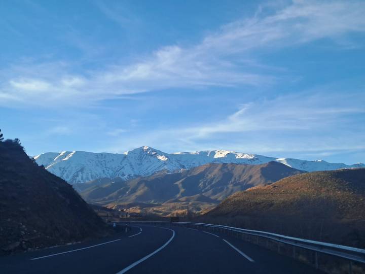 Snow-capped Atlas Mountains rising above rolling brown foothills and a curving highway at dawn light.