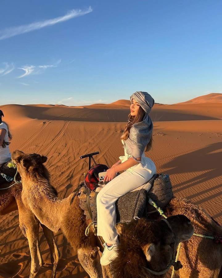 Young woman in traditional headscarf riding a camel across golden Sahara dunes at sunset.