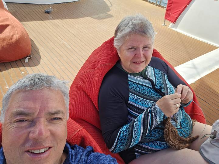 Selfie of two cruisers relaxing on deck chairs; woman knitting while man smiles at camera.