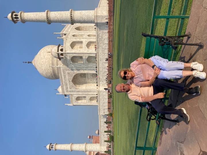 Smiling older couple sitting on a green bench with the Taj Mahal rising majestically behind them on a bright day