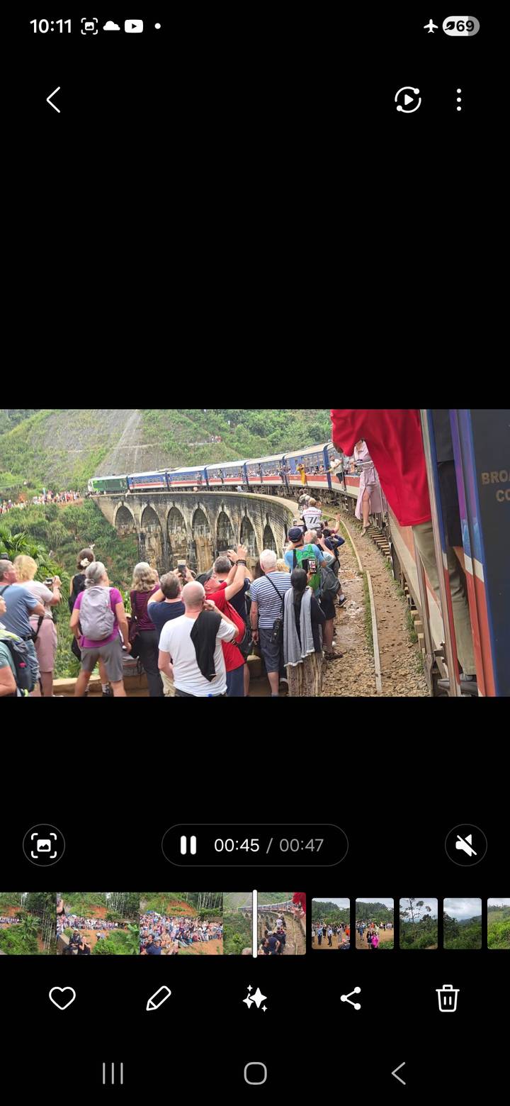 Tourists leaning out of a colorful passenger train as it crosses a stone viaduct in lush green hills.