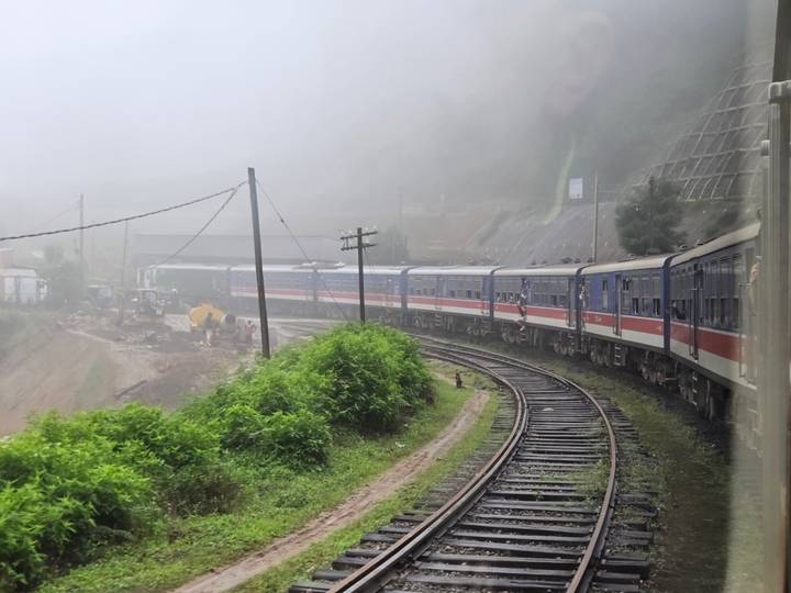 Long passenger train curving through misty green hills on a foggy day.