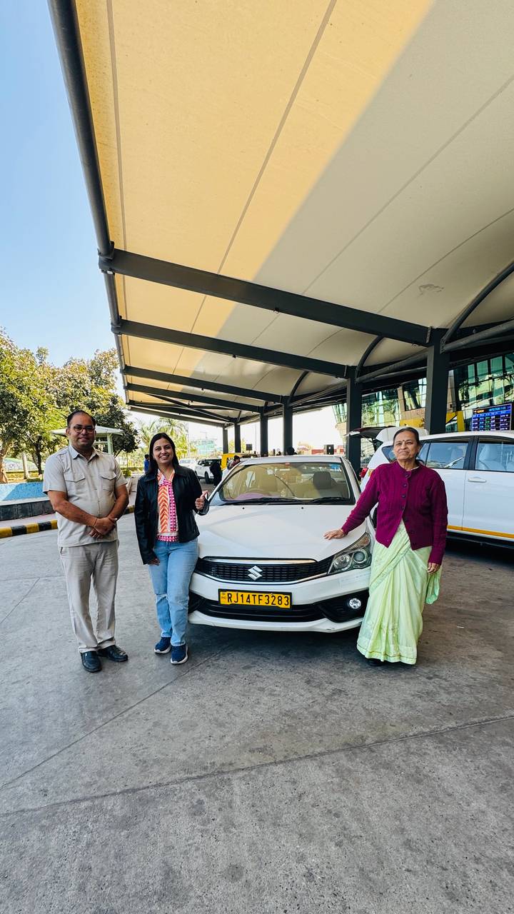 Tour guide, female traveller and elder woman posing in front of white Suzuki taxi outside Delhi airport