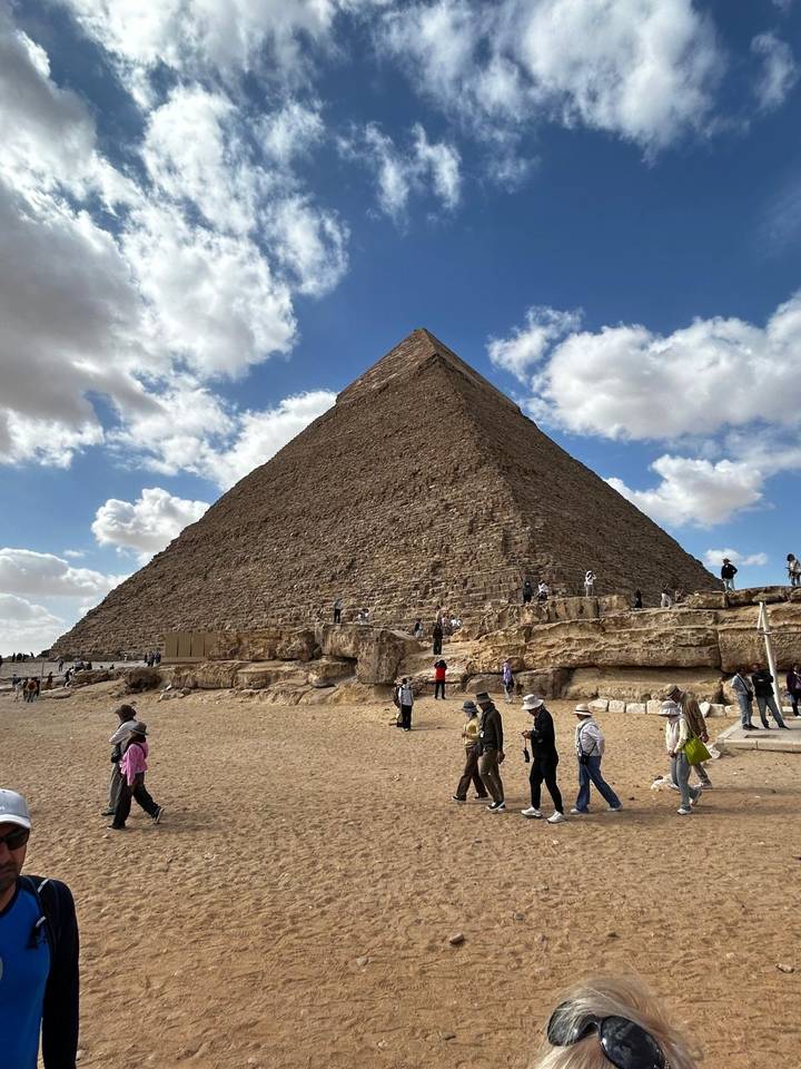 Iconic pyramid rising above sandy plateau dotted with tourists under a sky of cotton clouds.