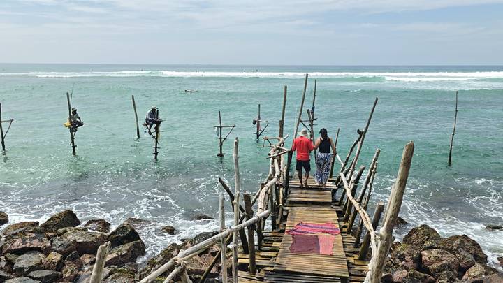 Tourists stand on wooden jetty watching traditional stilt fishermen over teal ocean waves.