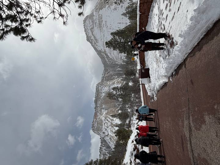 Visitors bundled in winter gear stand on a snowy roadside with layered cliffs and pines behind them.