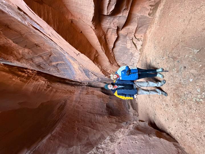 Two hikers pose inside a spacious red sandstone slot canyon with textured walls.