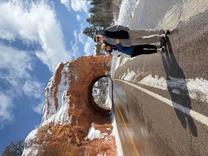 Couple stands on highway under a red rock tunnel dusted with snow and bright winter sunlight.