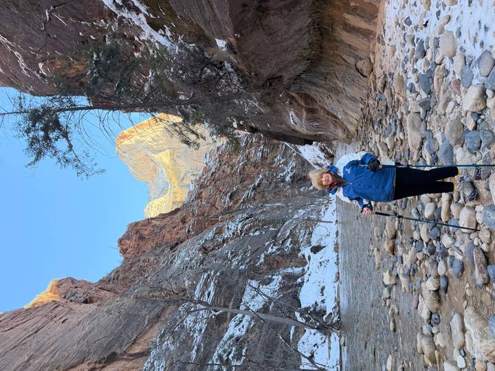 Hiker stands with poles on rocky riverbed between towering canyon walls in Zion’s Narrows.