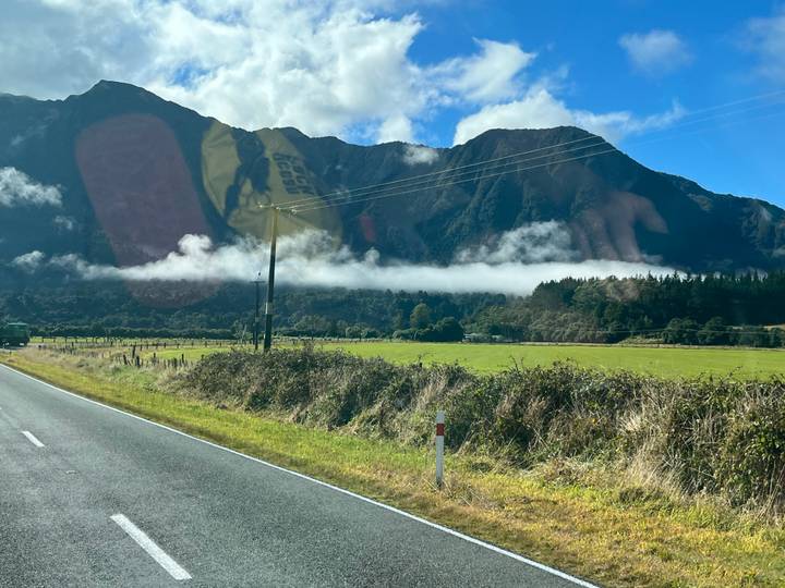 Country road leading toward mist-covered mountains with a ribbon of low cloud above green pastures.