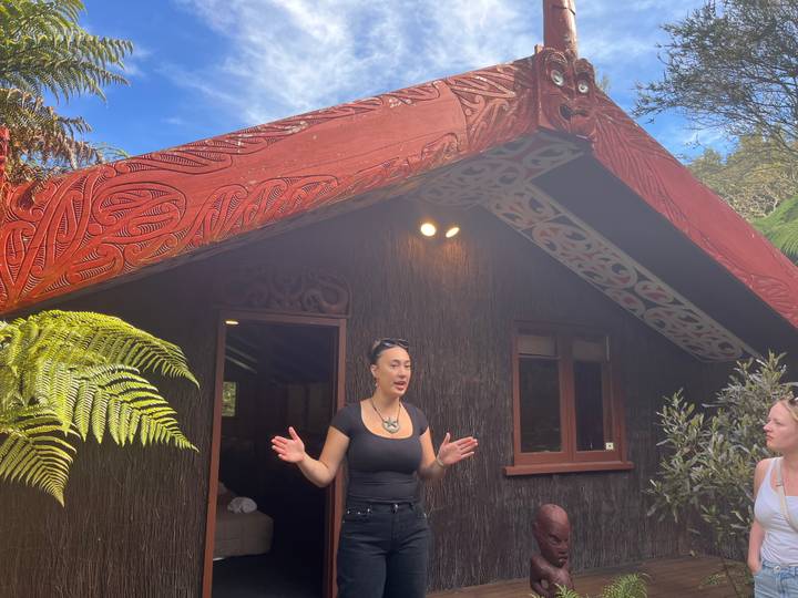 Maori guide speaks outside a carved meeting house while a visitor listens beside lush ferns.