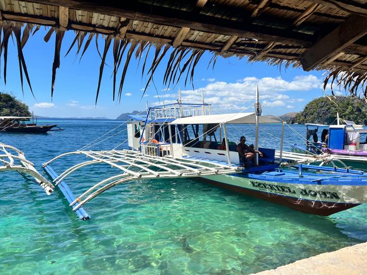 Traditional outrigger boat floating on clear turquoise water framed by a thatched roof, with distant islands on the horizon.