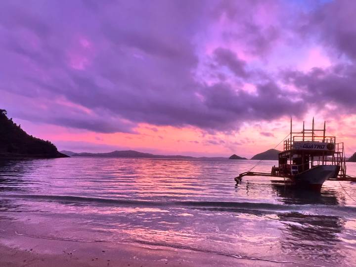 Silhouette of a wooden boat at anchor against a vivid purple-pink sunset over calm waters and island shapes.