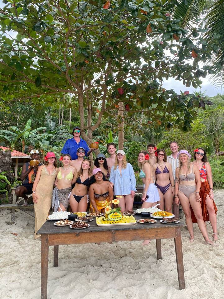 Large group of swimsuit-clad travellers posing under tropical trees on a beach holding coconuts