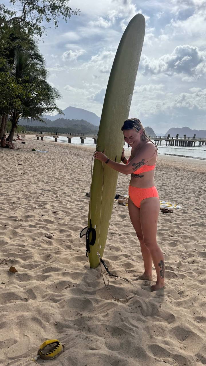 Woman in bright coral bikini standing on sandy beach holding a long surfboard near wooden pier