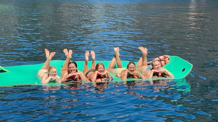 Five friends lying on a floating aqua mat, feet up, laughing on clear blue water