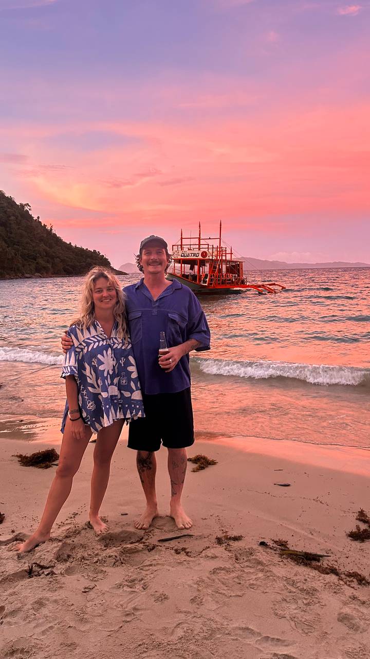 Smiling couple standing on a tropical beach at pastel sunset with a traditional outrigger boat anchored behind them