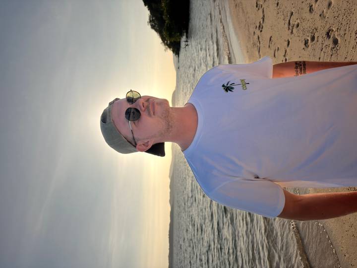 Young man in cap and sunglasses standing on a beach at sunset with the sun haloing behind his head and gentle waves beyond.