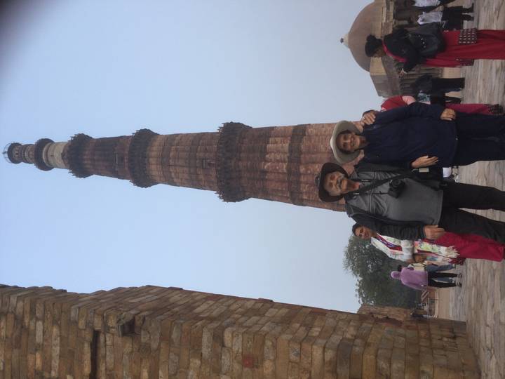 Elderly travellers stand before the soaring Qutub Minar while other visitors mill around.