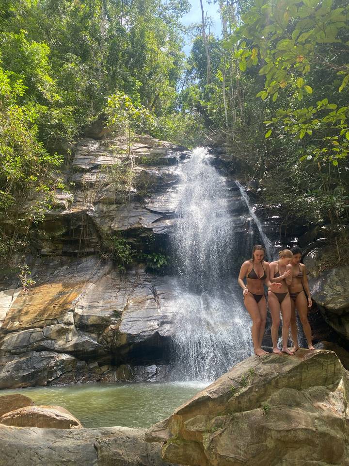 Three women in swimwear stand beneath a tropical waterfall in a lush jungle setting.