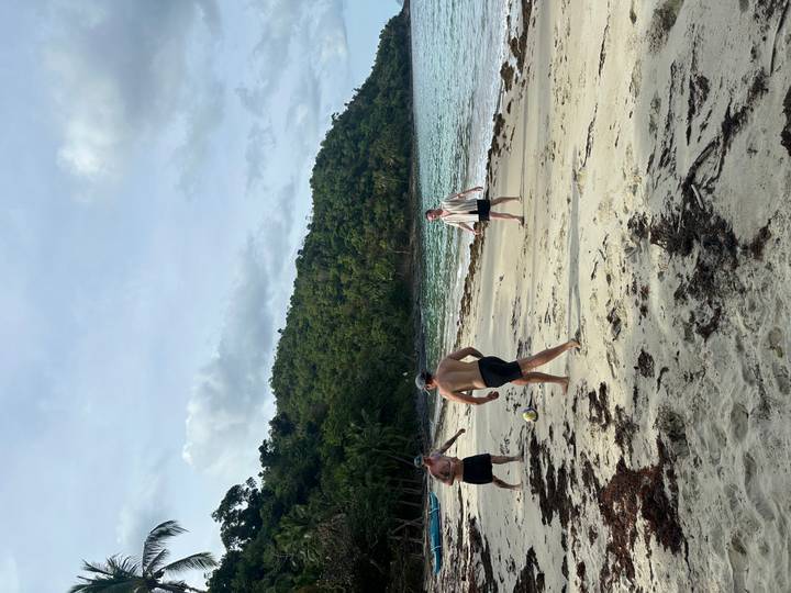 Three friends play football on a quiet tropical beach backed by green hillside.