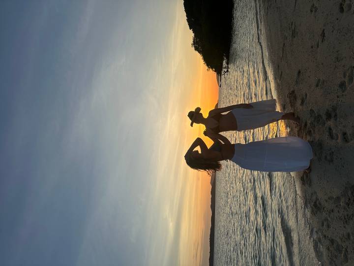 Silhouetted women in flowing skirts pose playfully on the shoreline at sunset.