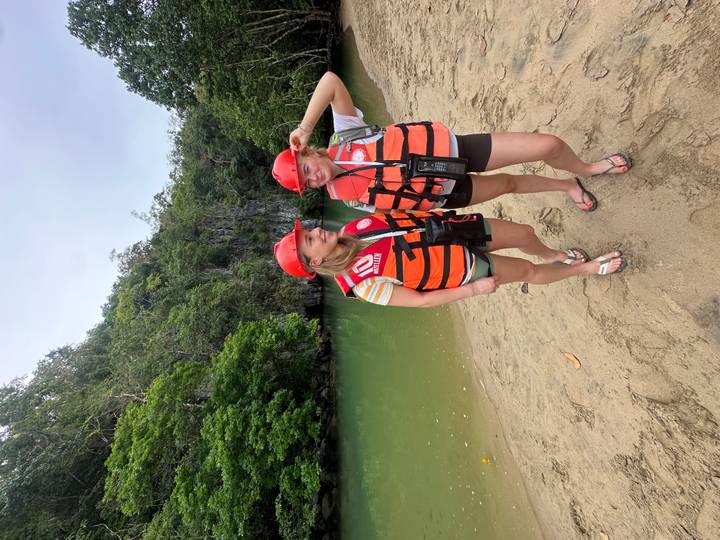 Two women wearing helmets and orange life vests stand on a sandy bank beside emerald water and lush cave entrance.