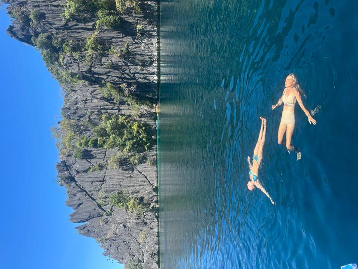 Two travelers float relaxed on their backs in a deep blue lagoon surrounded by jagged limestone cliffs.