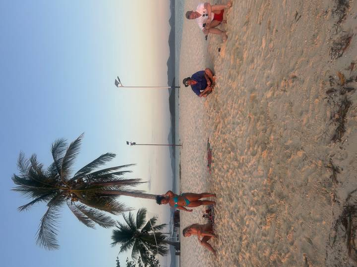 Sandy sunset beach with palm tree where travelers relax and a woman poses in turquoise swimwear.