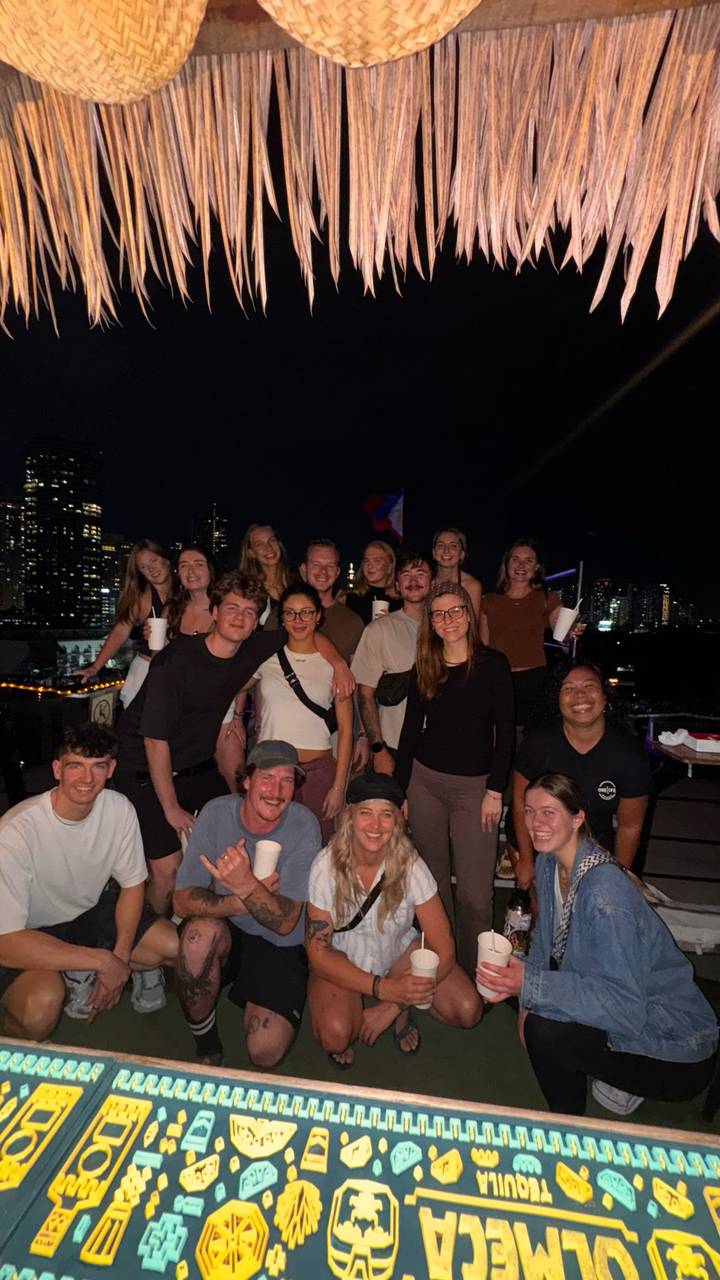 Large group of young travelers posing on a city rooftop at night with skyline behind.