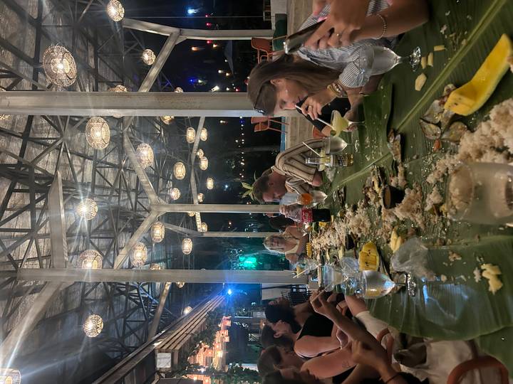 Long banana-leaf feast with travelers sharing food and drinks under decorative lights.