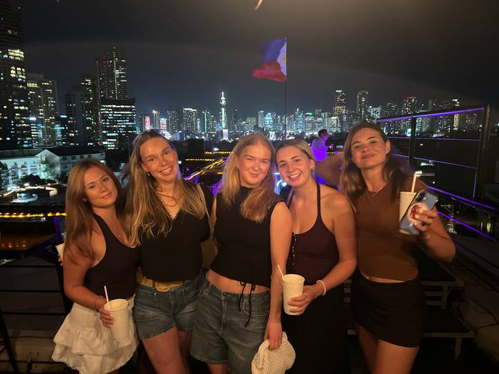 Five friends posing on a rooftop bar with a brightly lit city skyline behind them at night