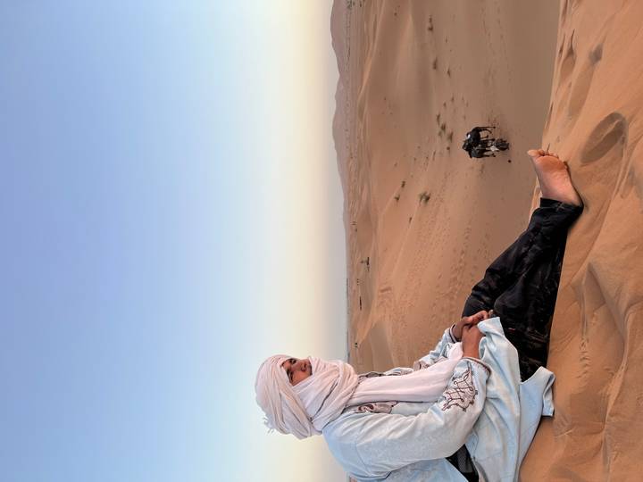 Berber man in turban sits on a sand dune overlooking vast desert with camel caravan below.