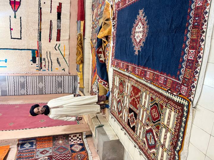 Berber carpet merchant standing among colourful traditional rugs displayed on the floor and walls.