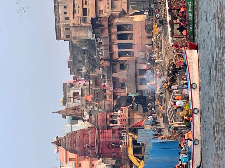 Busy riverbank ghats of Varanasi with cremation fires burning and colourful temples rising behind.